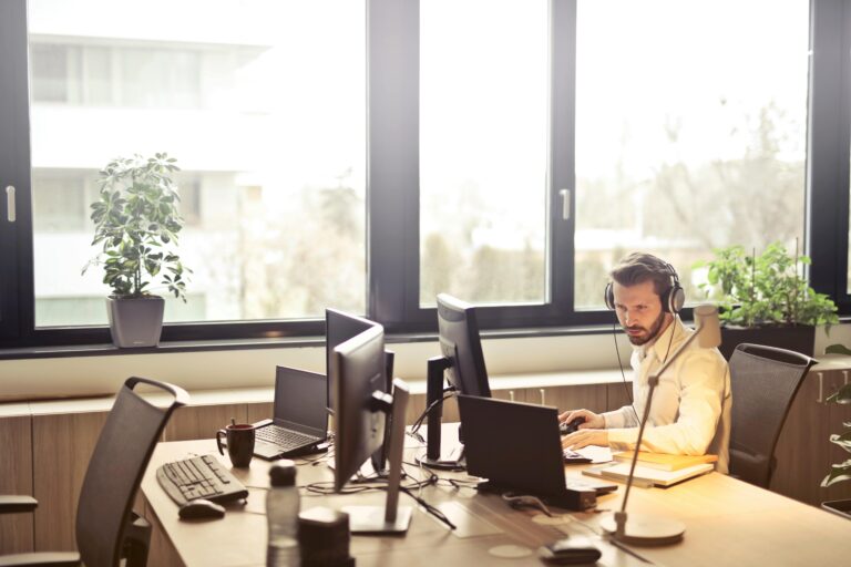 About Us A businessman sits at a desk using multiple computers and a headset in a well-lit modern office.